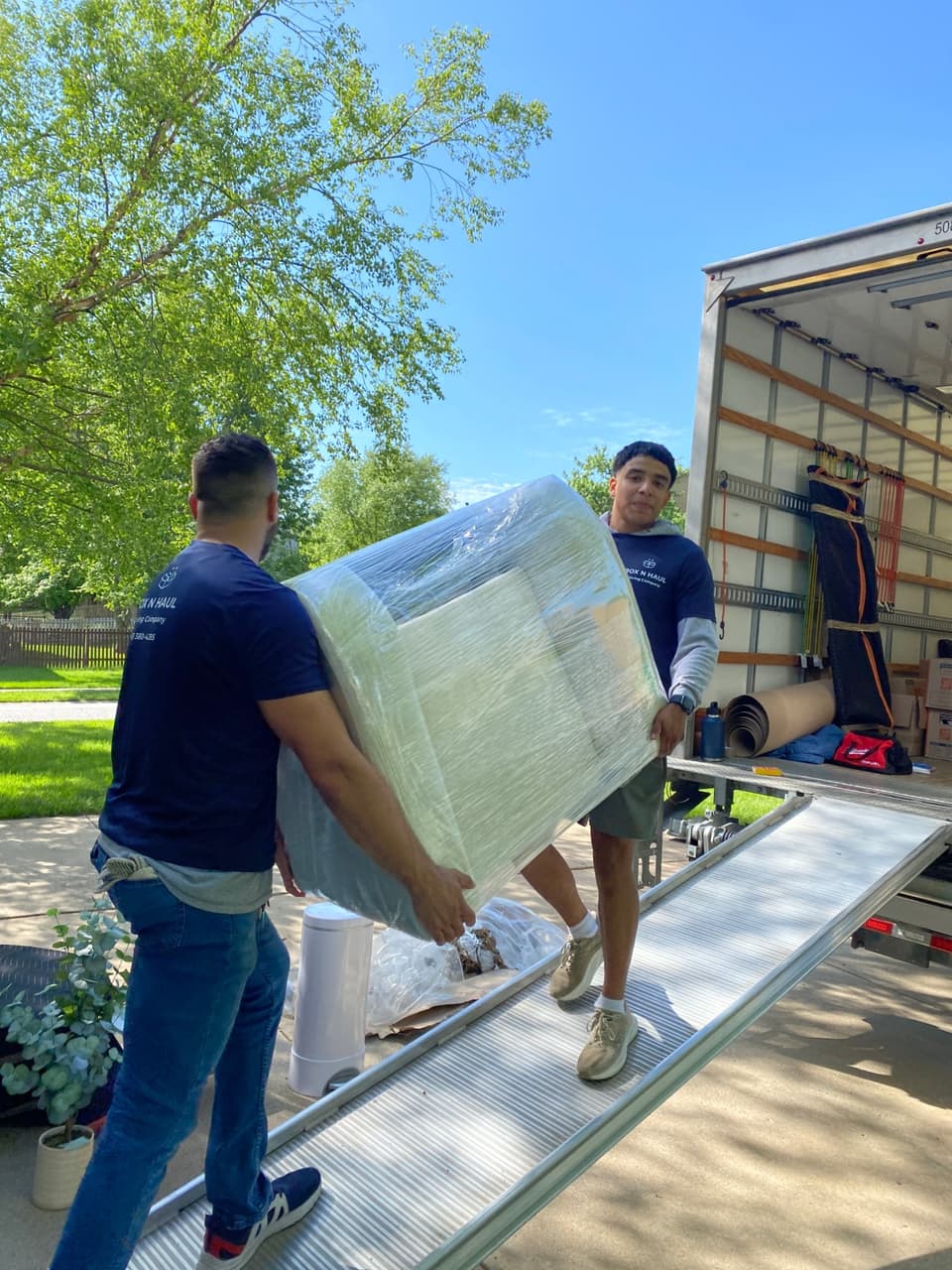 Two Box N Haul movers carrying a shrink-wrapped piece of furniture up the truck ramp on a sunny day.