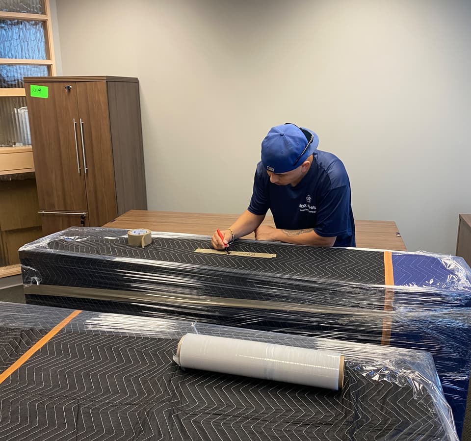 Box N Haul mover in a blue cap and branded shirt labeling shrink-wrapped office furniture with a marker before the move.