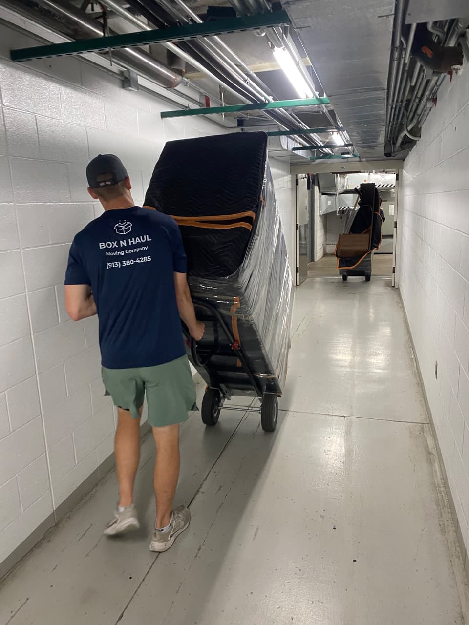 Box N Haul mover wheeling a tall, blanket-wrapped and shrink-wrapped item on a dolly down a building service corridor.