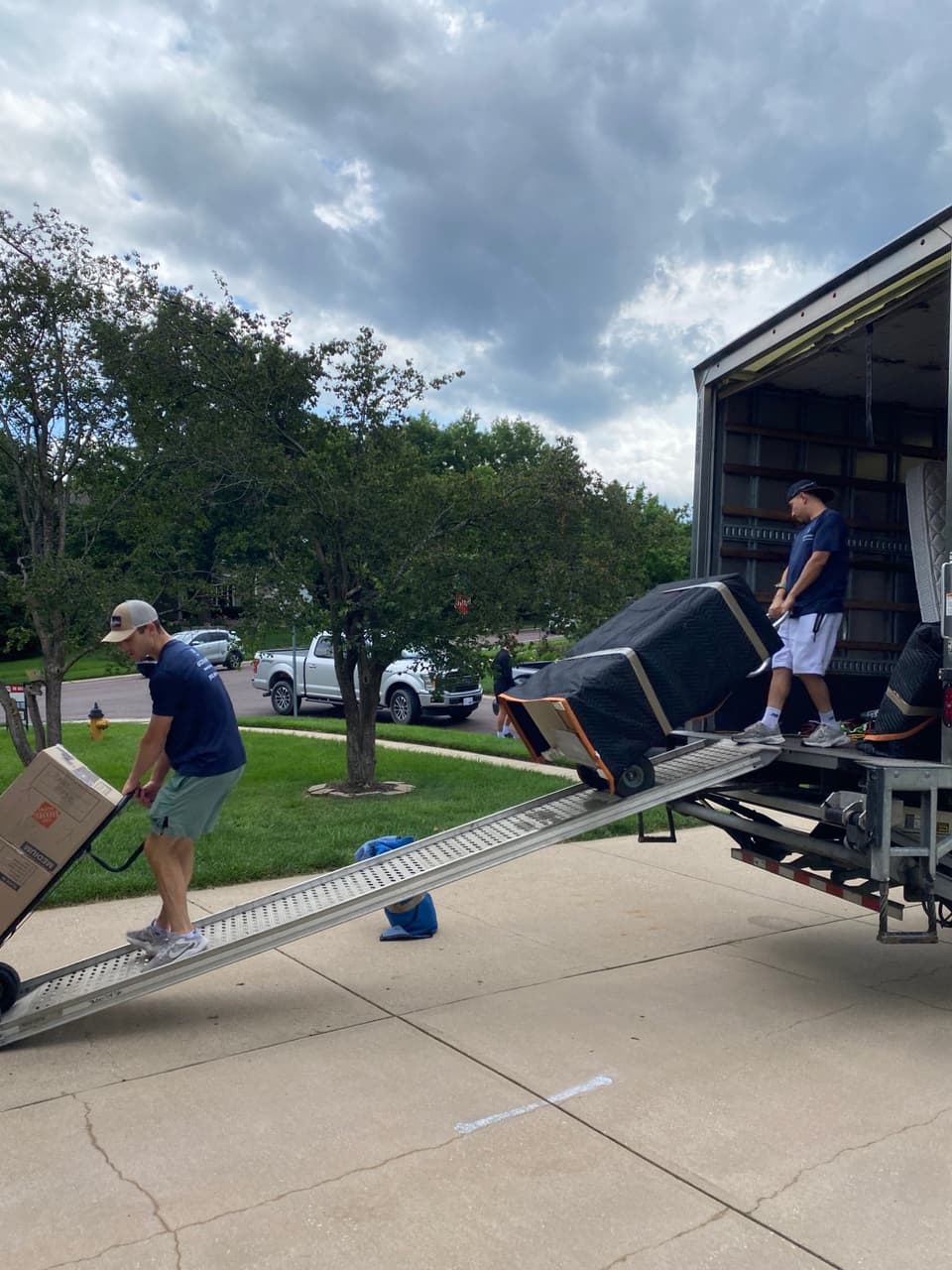 Two Box N Haul movers loading a blanket-wrapped item up the truck ramp on a hand truck while a teammate carries boxes.