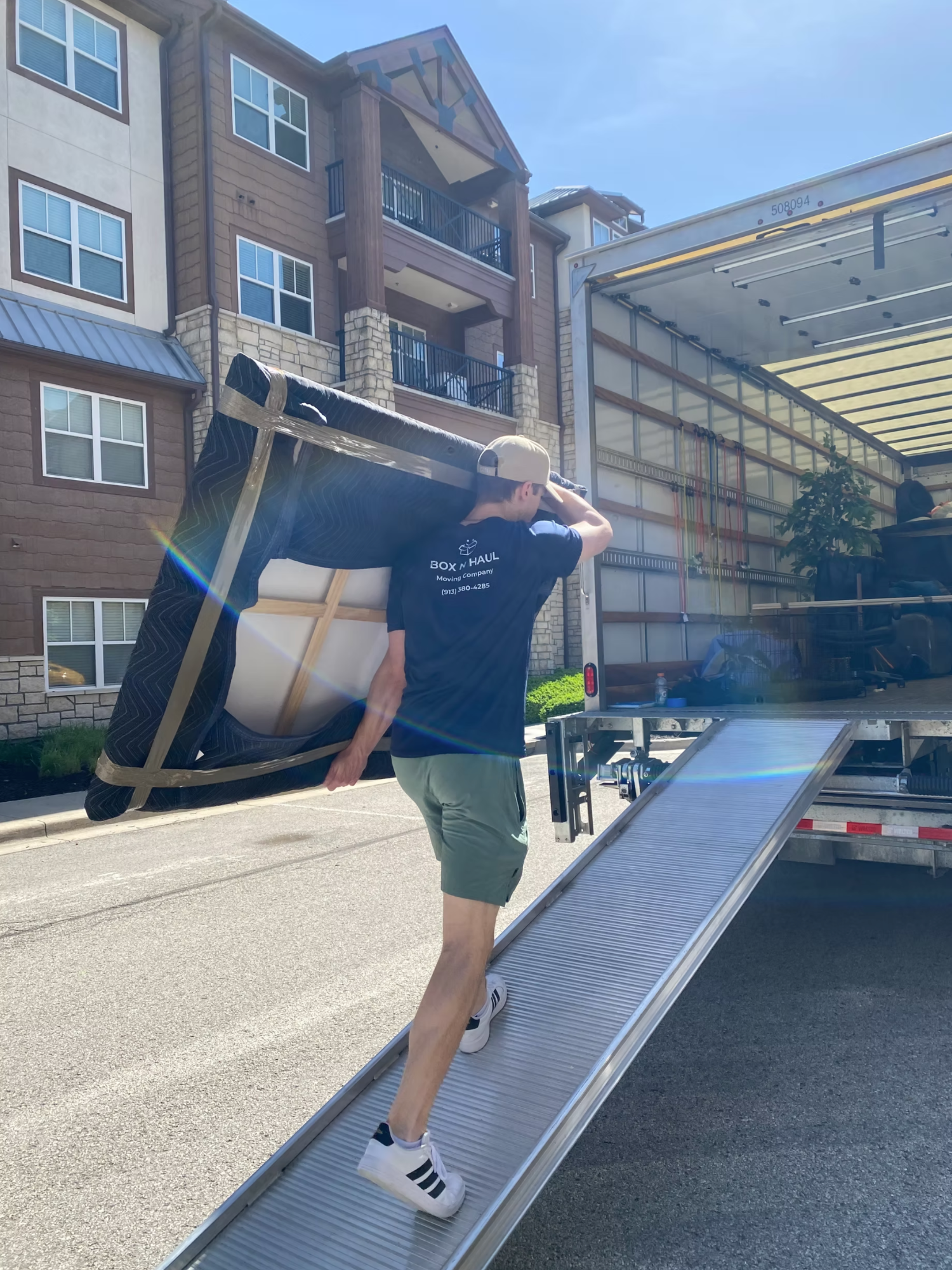 Box N Haul mover carrying a shrink-wrapped recliner up the ramp into the moving truck outside an apartment building.