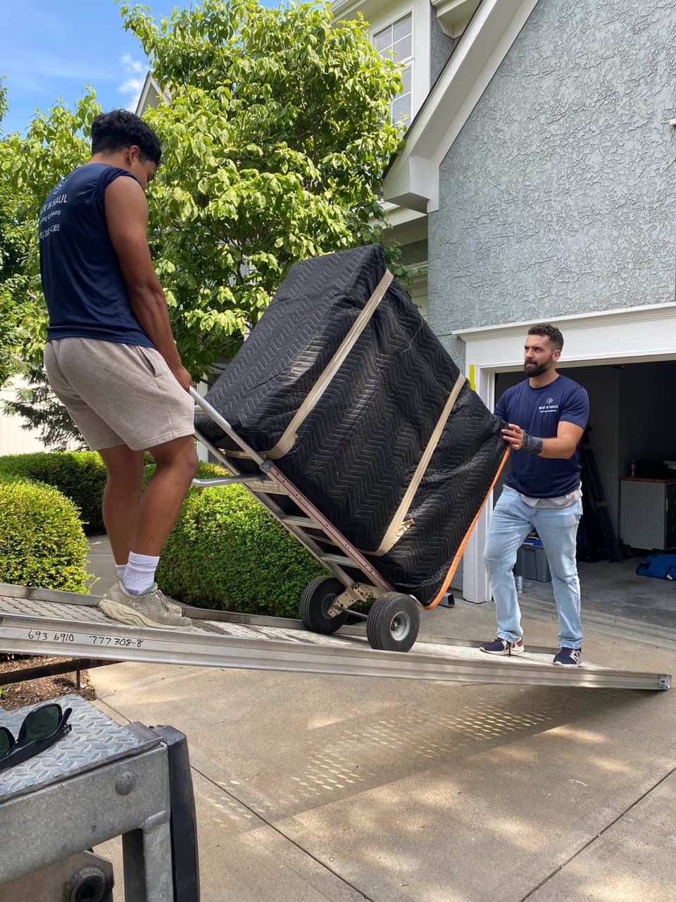 Box N Haul movers Cam and Alex wheeling a tall, blanket-wrapped piece of furniture down the truck ramp on a hand truck.