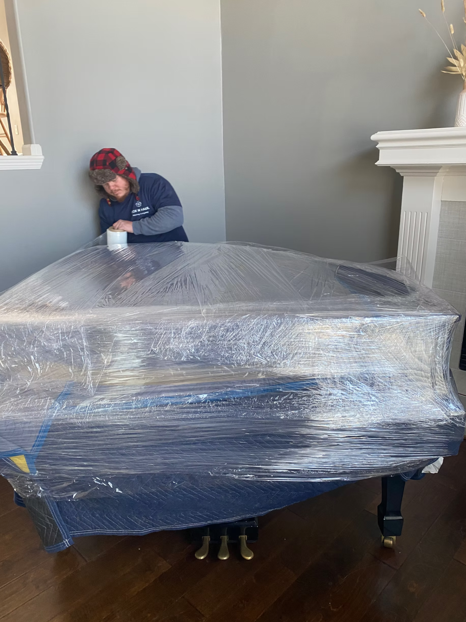 Box N Haul mover in a plaid trapper hat carefully shrink-wrapping a grand piano in a living room.