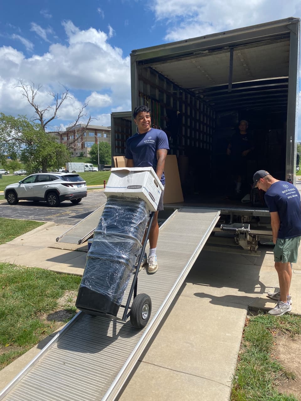 Smiling Box N Haul mover wheeling a Bankers Box and shrink-wrapped item up the truck ramp on a sunny day while a teammate assists.