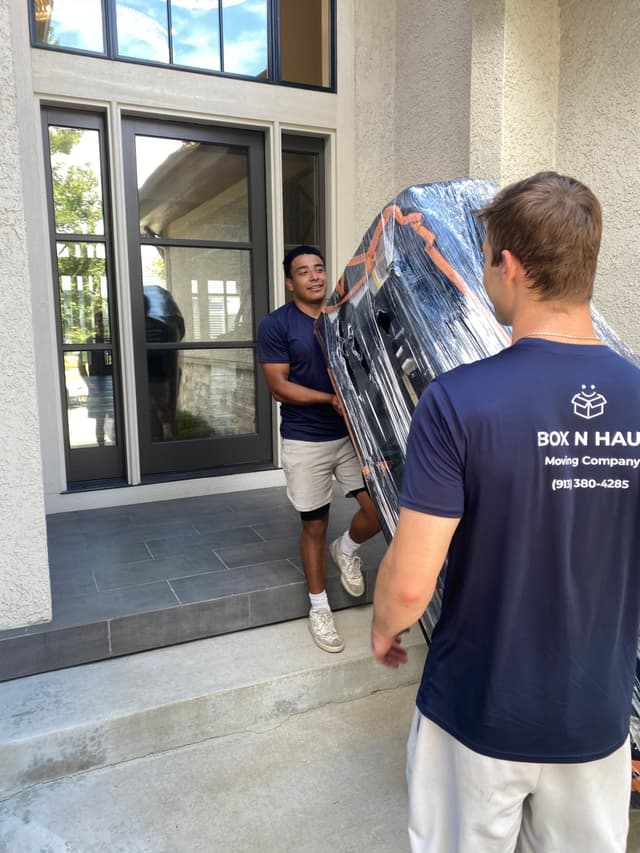 Two Box N Haul movers carrying a large shrink-wrapped piece of furniture through a glass front door.