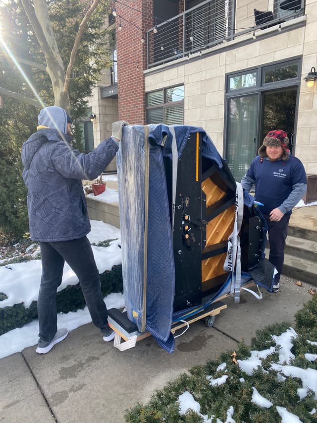 Two bundled-up Box N Haul movers wheeling a blanket-wrapped upright piano on a dolly up a snowy walkway on a bright winter day.