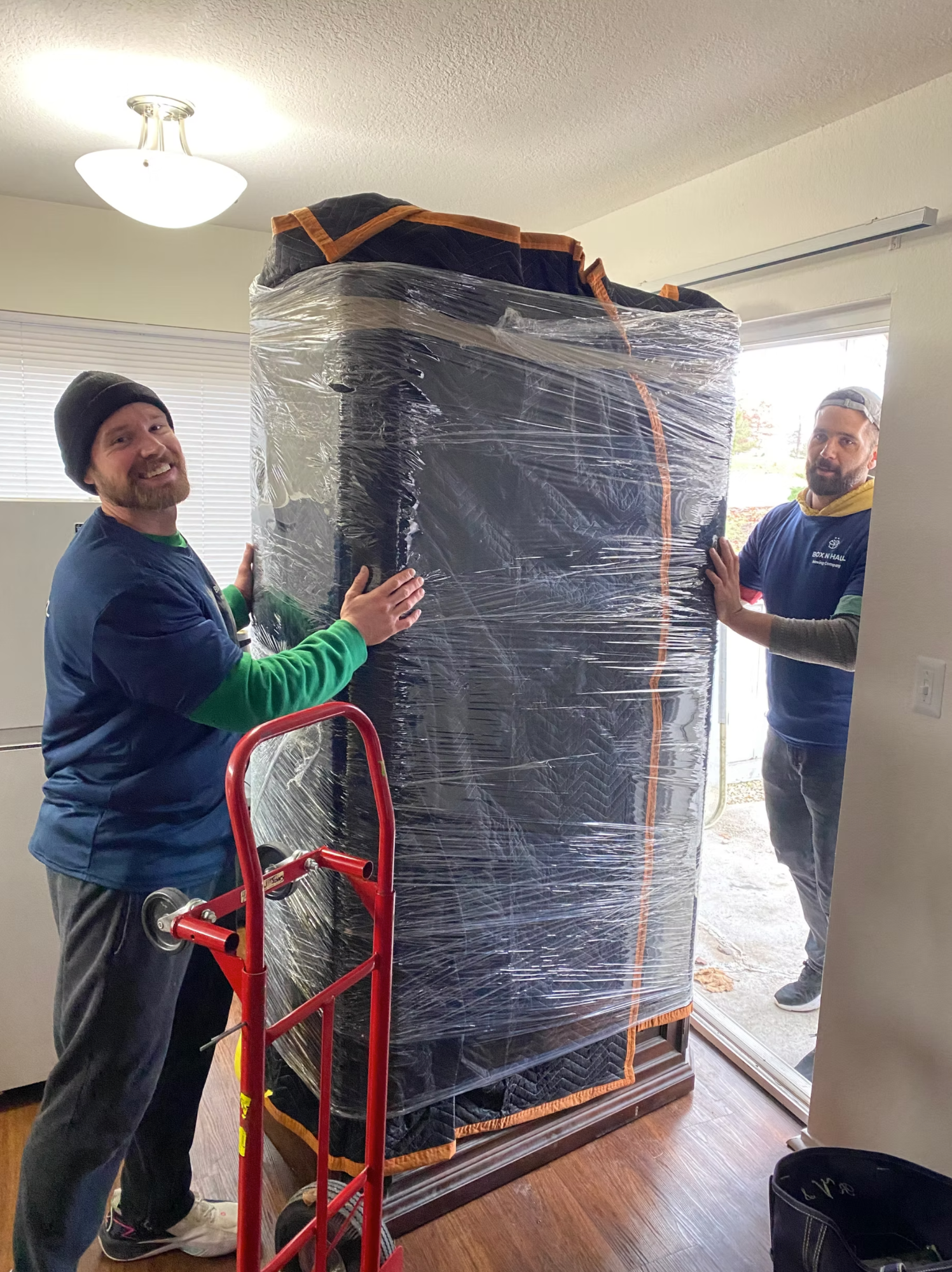 Two smiling Box N Haul movers wheeling a tall, blanket-wrapped piece of furniture through a doorway on a dolly.