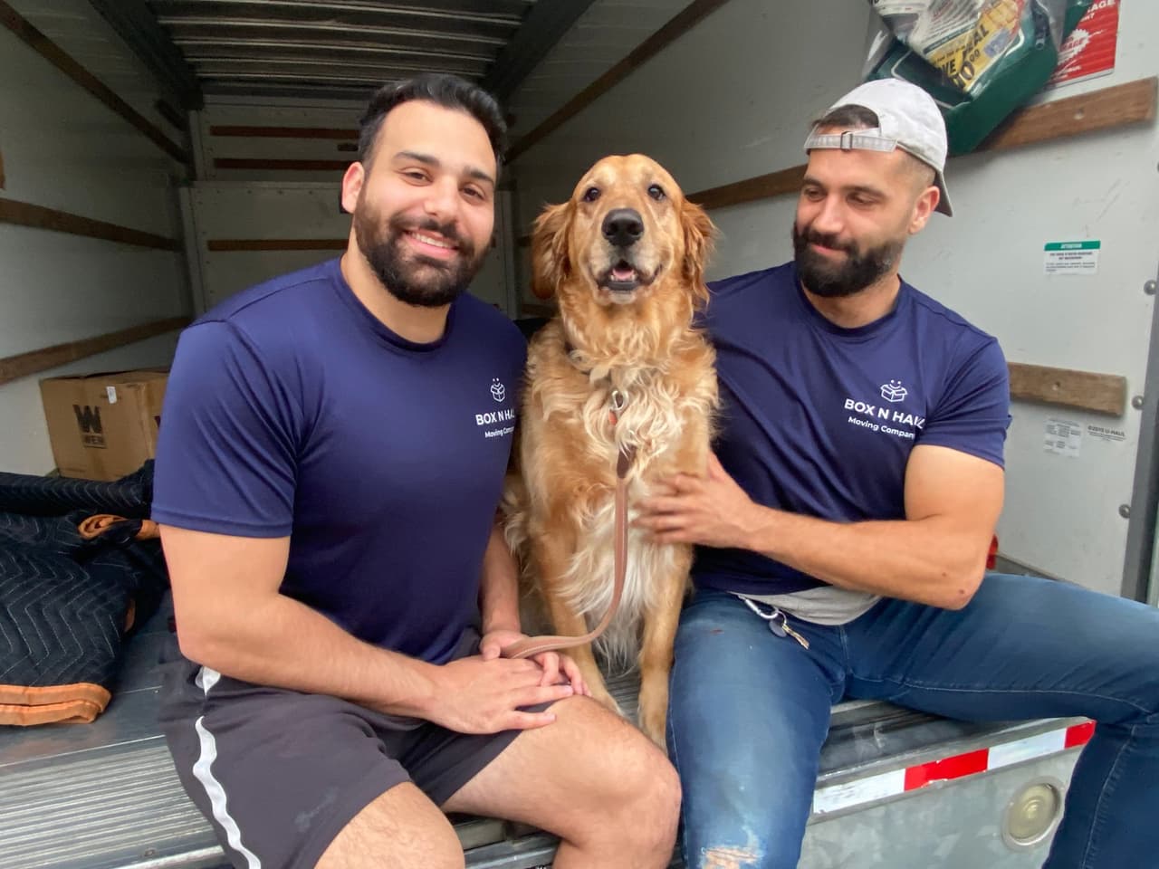 Two smiling Box N Haul movers sitting in the back of a moving truck with a golden retriever.